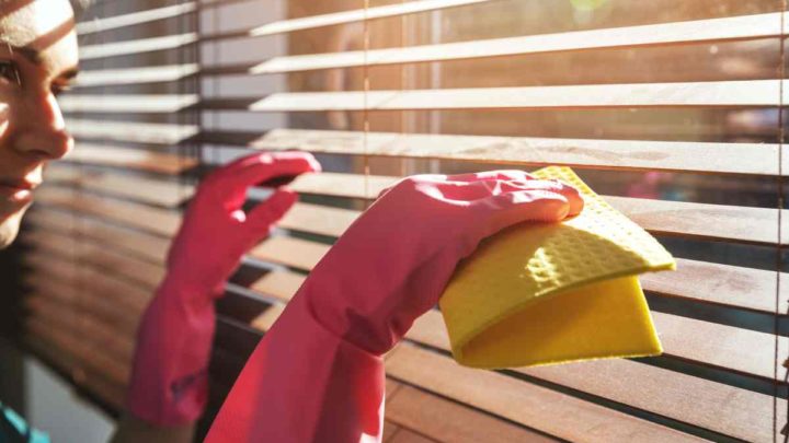 A person wearing pink cleaning gloves is cleaning dusty wood blinds with a yellow rag at White House Designs For Life near New Jersey (NJ)