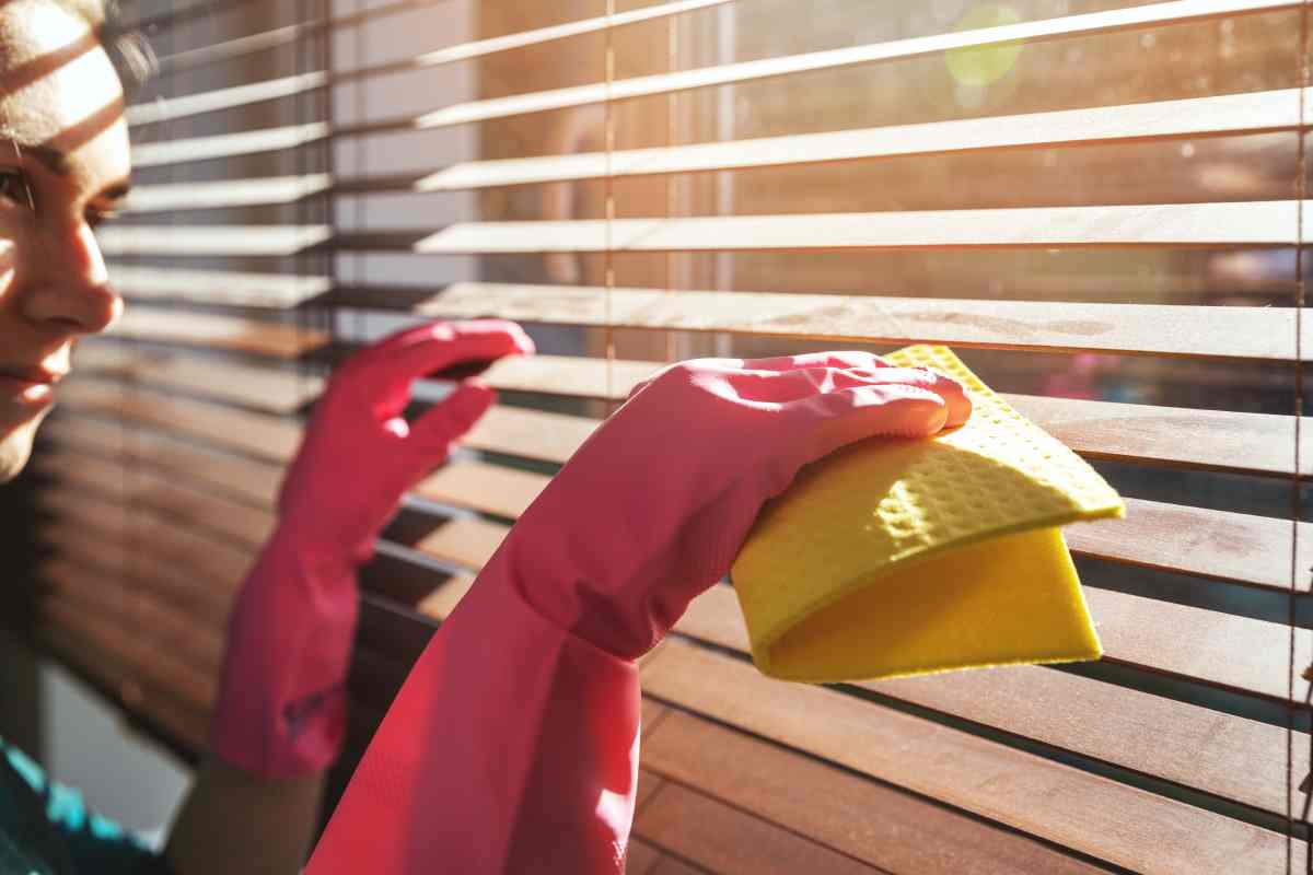 A person wearing pink cleaning gloves is cleaning dusty wood blinds with a yellow rag